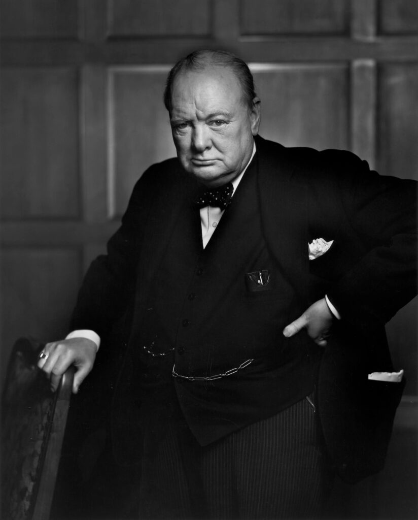 Churchill, 67, wearing a suit, standing and holding into the back of a chair